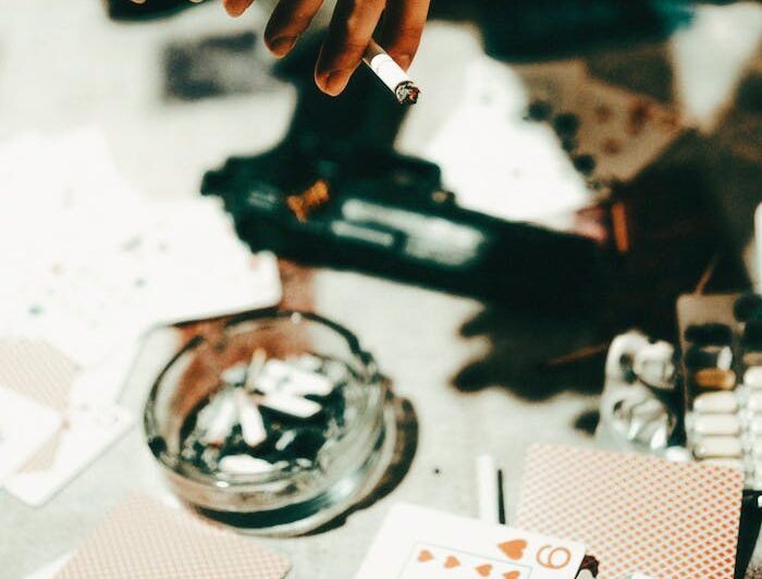 Close-up shot showing hands holding a cigarette near scattered playing cards and a pistol on a table.
