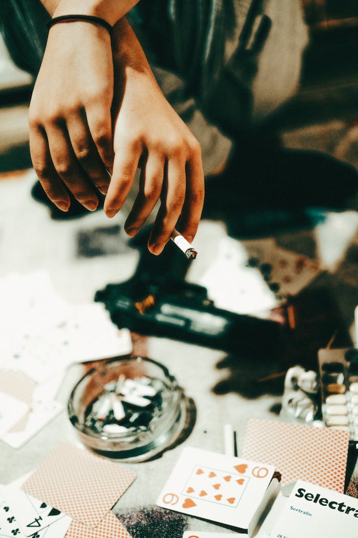 Close-up shot showing hands holding a cigarette near scattered playing cards and a pistol on a table.
